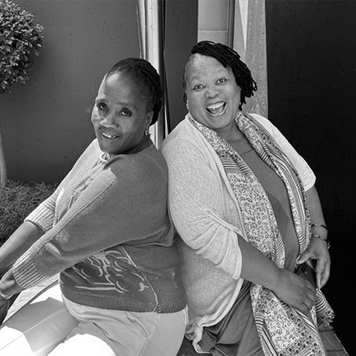 Two women sitting back-to-back outdoors, smiling warmly at the camera.